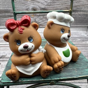 May include: Two brown bear figurines sitting side by side. The left bear wears a red bow on its head and a white apron. The right bear wears a white chef hat and a white apron with a green accent. Both bears have blue eyes and white muzzles.