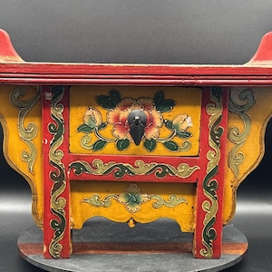 May include: A small wooden chest with a red top featuring curved ends, a yellow body, and a single drawer. The front is decorated with hand-painted floral motifs in red, green, and yellow, and includes a black metal pull handle on the drawer.