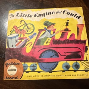 May include: Vintage children's record album cover featuring a red train with a conductor, a bicycle, and other toys. The title "The Little Engine That Could" is displayed in large letters against a yellow background. The Golden Record logo is in the lower left corner.