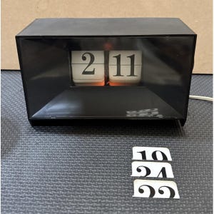 May include: A black retro flip clock displaying the time as 2:11. The clock has white numbers on a black background. Additional number cards are below the clock.