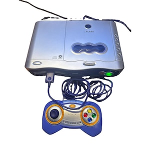 May include: A silver V.Flash video game console with a blue and silver controller connected by a cord. The console features a disc drive, a power button with a green light, and the V.Flash logo. The controller has a joystick, four coloured buttons, and a central orange button.