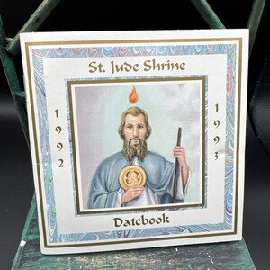 May include: A square 1992-1993 St. Jude Shrine datebook featuring a central illustration of a bearded figure in a blue cloak holding a staff and a gold medallion, with a flame above the head, framed by a blue and white marbled border.
