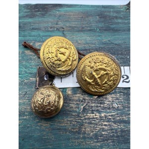 May include: Three antique gold-coloured buttons with detailed nautical designs. Two larger buttons and one smaller button are visible. The buttons feature anchors, wreaths, and intricate detailing. A measuring tape is partially visible.