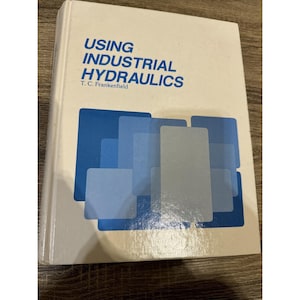 May include: A white book titled "USING INDUSTRIAL HYDRAULICS" with blue and grey geometric shapes on the cover. The author's name, T. C. Frankenfield, is printed below the title. The book shows signs of wear.
