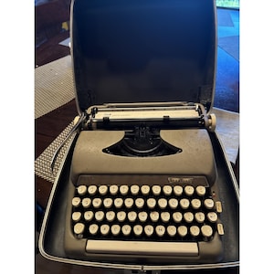 Puede incluir: Una máquina de escribir Sterling vintage en su estuche. La máquina es gris con teclas blancas y una cinta negra. El estuche está abierto, revelando la máquina. Las teclas están en disposición QWERTY. Se ve la palabra 'STERLING'.