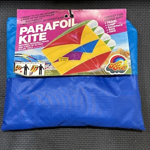 Puede incluir: Un cometa Parafoil azul en su embalaje. El embalaje presenta un diseño de cometa colorido con secciones rojas, amarillas y azules. El texto en el embalaje incluye "Parafoil Kite", "Ready to Fly" y "Perfect for People Who: Travel, Camp, Backpack, Bike, Hike."