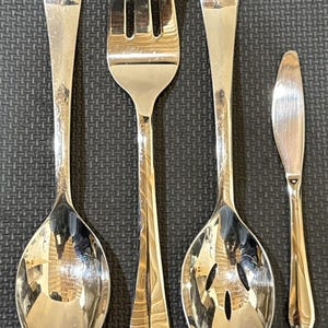 May include: A set of four silver-toned stainless steel utensils consisting of a solid spoon, a three-tined fork, a slotted spoon with three oval openings, and a small butter knife, all featuring smooth, reflective surfaces and rounded handles.