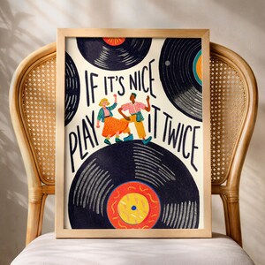 Könnte beinhalten: Ein gerahmter Kunstdruck mit einer skurrilen Illustration eines tanzenden Paares vor dem Hintergrund von Schallplatten. Der Druck enthält den Text "If it's nice play it twice". Das Kunstwerk befindet sich in einem Holzrahmen und wird auf einem Korbstuhl ausgestellt.