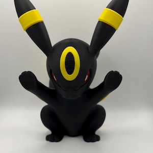 May include: A black figurine of a creature with long pointed ears, featuring bright yellow bands on the ears and a yellow oval marking on the forehead. The creature has red eyes and is posed with both front paws raised in a forward-facing position.