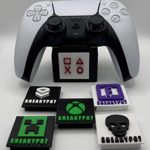 May include: A white and black PlayStation 5 controller with a black stand featuring red, white, and black symbols. Below are six controller button covers with various logos and the text "SNEAKYP87".