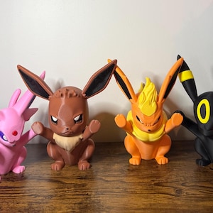 May include: A set of four stylized character figurines featuring Espeon in pink, Eevee in brown, Flareon in orange and yellow, and Umbreon in black with yellow rings. Each figure is depicted in a dynamic pose with raised arms and pointed ears.