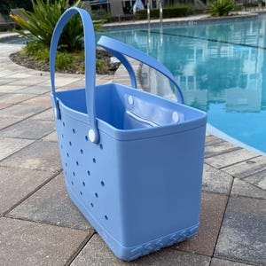 May include: A light blue tote bag with a rectangular shape and a handle. The bag has a series of small, evenly spaced holes on the sides. The bag is made of a durable material and is suitable for carrying items. The bag is next to a swimming pool.