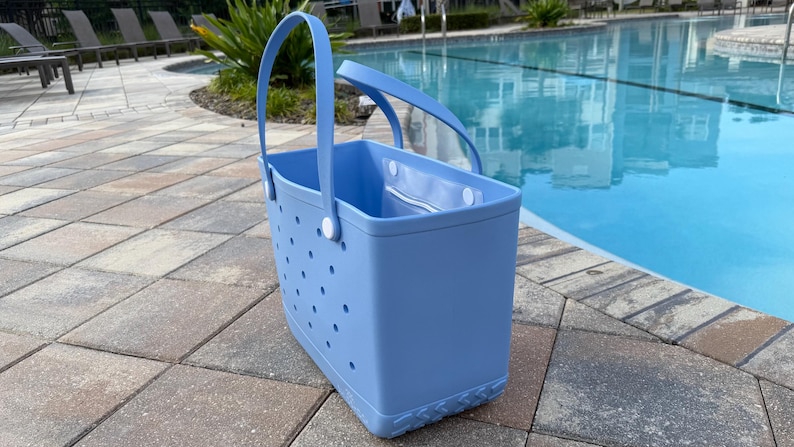 May include: A light blue tote bag with a rectangular shape and a handle. The bag has a series of small, evenly spaced holes on the sides. The bag is made of a durable material and is suitable for carrying items. The bag is next to a swimming pool.