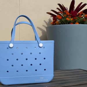 May include: A light blue tote bag with a rectangular shape and a perforated design. The bag has two handles and white fasteners. A large blue planter with orange flowers and dark red leaves is in the background.