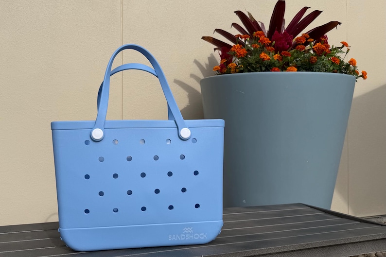May include: A light blue tote bag with a rectangular shape and a perforated design. The bag has two handles and white fasteners. A large blue planter with orange flowers and dark red leaves is in the background.