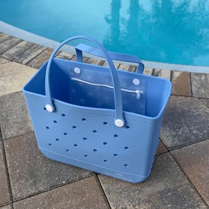May include: A light blue tote bag with a rectangular shape and two handles. The bag has a series of small, evenly spaced holes and a zippered pocket. The bag is sitting on a stone surface next to a pool.