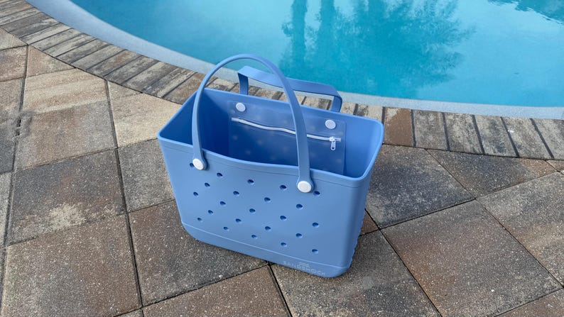 May include: A light blue tote bag with a rectangular shape and two handles. The bag has a series of small, evenly spaced holes and a zippered pocket. The bag is sitting on a stone surface next to a pool.