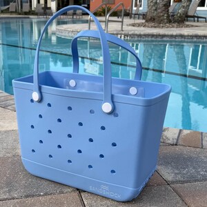 May include: A light blue tote bag with two handles and a rectangular shape. The bag has numerous small, round holes and white fasteners. The bag is sitting on a stone surface next to a swimming pool.