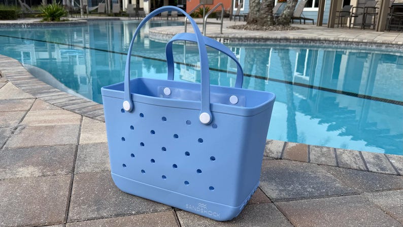 May include: A light blue tote bag with two handles and a rectangular shape. The bag has numerous small, round holes and white fasteners. The bag is sitting on a stone surface next to a swimming pool.