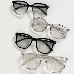 Puede incluir: Se muestran cuatro pares de gafas con lentes transparentes. Dos pares tienen monturas negras y los otros dos tienen monturas transparentes. Las gafas tienen forma cuadrada y están dispuestas sobre una superficie blanca.