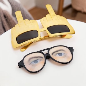 May include: Two pairs of novelty sunglasses. One pair is gold-coloured and shaped like hands giving the middle finger, with black lenses. The other pair is black-framed glasses with printed eyes.