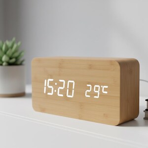 May include: A rectangular wooden digital alarm clock displaying the time as 15:20 and the temperature as 29°C. The clock has a light-colored wood grain finish and a white LED display. A small plant in a white pot is visible in the background.