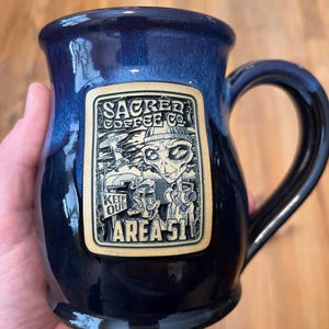 May include: A blue ceramic coffee mug with a black handle and a rectangular graphic. The graphic features an alien with the text "Sacred Coffee Co." and "Area 51". The mug has a glossy finish.