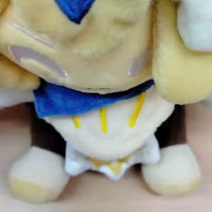 May include: A plush toy in a seated position, featuring a yellow body with a blue diamond-shaped emblem. The toy has a blue collar, white accents, and brown legs with cream-colored feet. The design includes yellow vertical stripes.