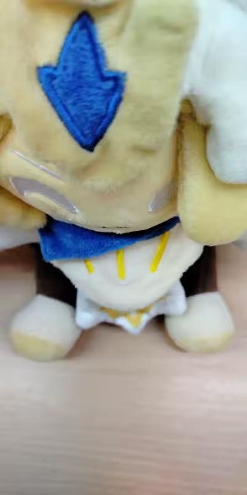 May include: A plush toy in a seated position, featuring a yellow body with a blue diamond-shaped emblem. The toy has a blue collar, white accents, and brown legs with cream-colored feet. The design includes yellow vertical stripes.