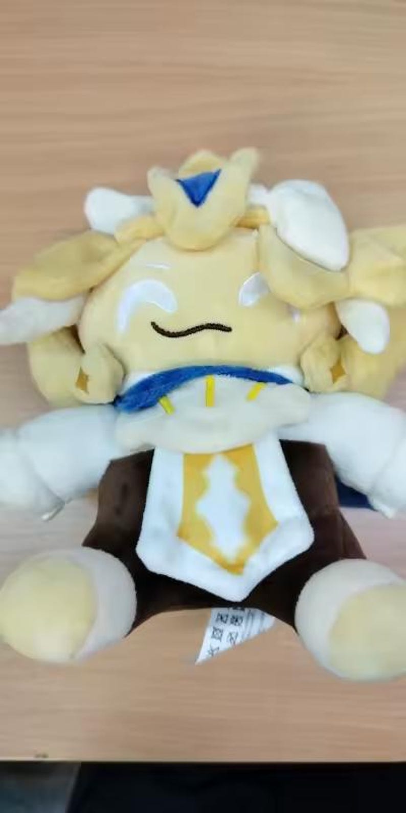 May include: A plush toy character with a yellow head, white arms, and a brown torso. The character has a blue collar and a yellow and white emblem on its chest. The toy is sitting on a wooden surface.