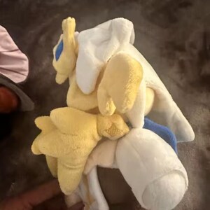 May include: A plush toy in yellow, white, and blue. The toy has a rounded shape with multiple soft, stuffed appendages. The plushie appears to be a character from a video game or cartoon.