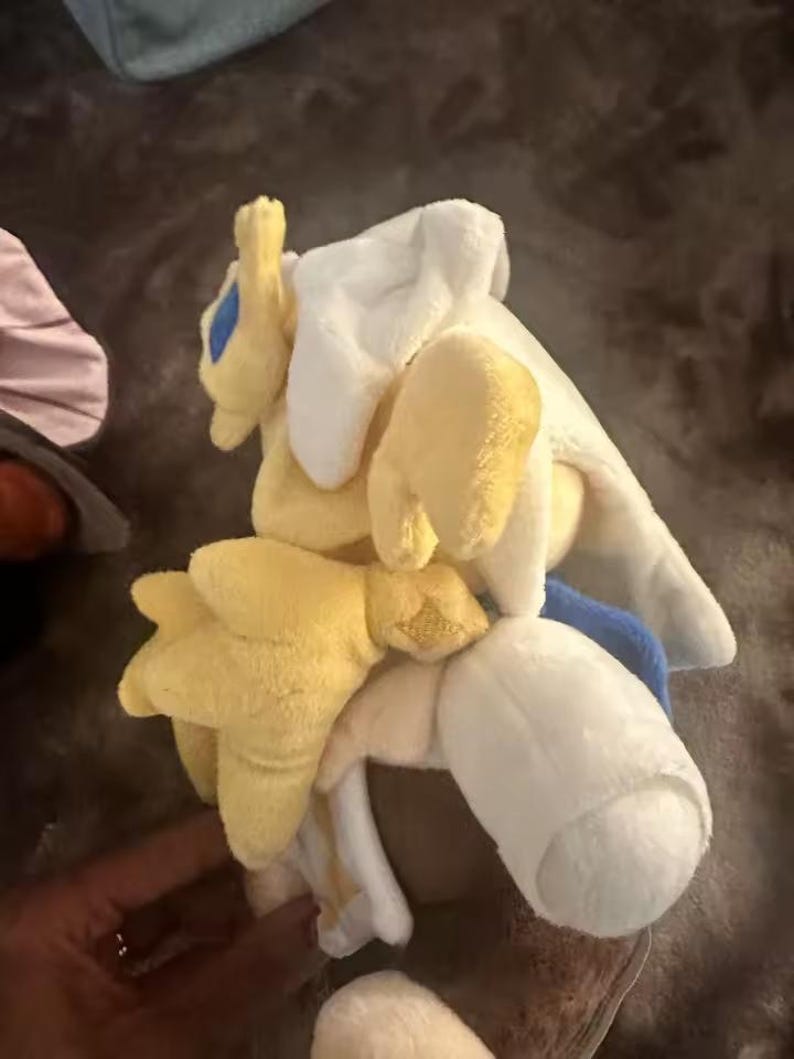 May include: A plush toy in yellow, white, and blue. The toy has a rounded shape with multiple soft, stuffed appendages. The plushie appears to be a character from a video game or cartoon.
