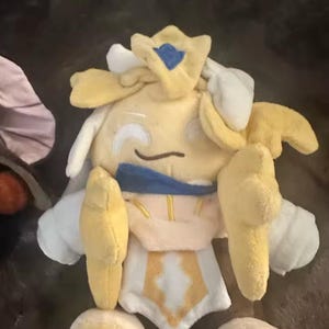 May include: A plush toy in the shape of a character with a yellow body, white accents, and a blue crown. The toy has a smiling face and is made of soft fabric. The character has yellow arms and legs.