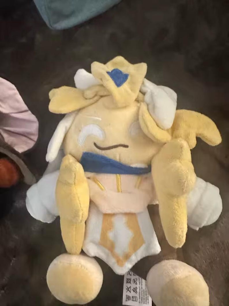 May include: A plush toy in the shape of a character with a yellow body, white accents, and a blue crown. The toy has a smiling face and is made of soft fabric. The character has yellow arms and legs.