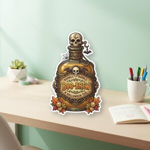 Peut inclure: Autocollant sur le thème d'Halloween avec une bouteille de potion dorée. Elle est ornée d'un bouchon en forme de crâne et d'une étiquette indiquant "Double Bubble Boo-Brew & Halloween Toil". Des feuilles décoratives et une chauve-souris sont également présentes.