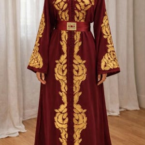 May include: A burgundy-colored, long-sleeved dress with gold embroidery. The dress features ornate gold patterns on the sleeves, bodice, and along the front. A matching burgundy belt with a gold buckle cinches the waist. The dress is floor-length.