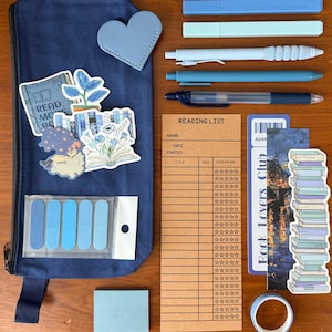May include: A dark blue pencil case decorated with various stickers, including a book, a plant, and a character. Beside it are blue pens, highlighters, a brown reading list notepad, a bookmark labeled Book Lovers Club, and a roll of blue tape.
