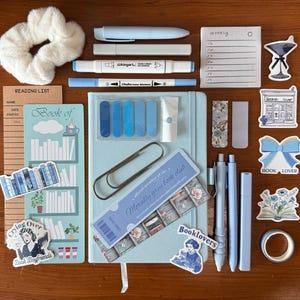 May include: A collection of blue and white stationery items, including pens, markers, stickers, a notebook, and a scrunchie. Decorative stickers feature the text "Booklovers" and "Bookish Lover". The items are arranged to suggest a theme of reading and book clubs.