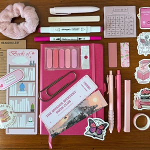 Puede incluir: Una colección de artículos de papelería rosas, que incluyen un diario, bolígrafos, resaltadores, notas adhesivas, pegatinas y una scrunchie, dispuestos sobre una superficie de madera. Los artículos tienen texto que dice "Book of", "Small Town Romances", "The Murder Mystery Book Club" y "Books are my language".
