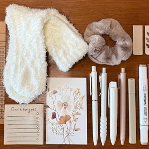 Puede incluir: Una colección de artículos de papelería y accesorios, que incluyen bolígrafos, pegatinas, una goma para el pelo y una bufanda esponjosa. Los artículos están dispuestos sobre una superficie de madera. También hay plantillas de listas de lectura y un pequeño bloc de notas con el texto "¡No te olvides!"