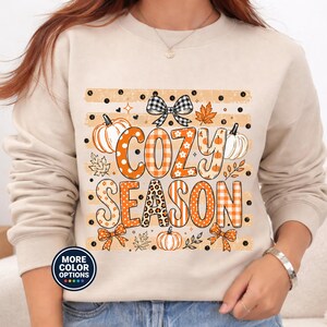 Peut inclure: Un sweat-shirt à col rond beige avec une impression graphique portant le texte COZY SEASON dans des motifs orange et blancs. Le design comprend des illustrations de citrouilles, de feuilles d'automne, de nœuds à carreaux noirs et blancs.