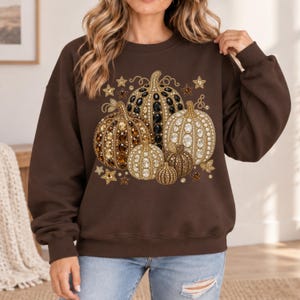 May include: A dark brown crewneck sweatshirt featuring a central graphic of five pumpkins decorated with various brown, gold, and black faceted gems and pearls. The design is accented with small gold stars and swirling vine details.