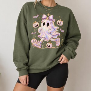 May include: An olive green crewneck sweatshirt featuring a central graphic of a holographic ghost wearing a purple bow and sneakers, surrounded by small purple bats, smiling light purple pumpkins, and gold stars against the dark fabric.