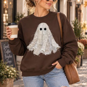 May include: A dark brown crewneck sweatshirt featuring a large, central ghost graphic constructed from various off-white fabric patches, including lace and textured textiles, with two solid black circular eyes and frayed edges.