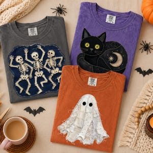 Peut inclure: Trois t-shirts avec des motifs d'Halloween : un t-shirt gris anthracite avec trois squelettes dansants, un t-shirt violet avec un chat noir et une lune, et un t-shirt orange avec un fantôme blanc. Chaque t-shirt a un col rond et des manches courtes.