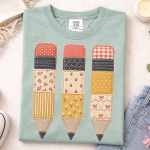 May include: A sage green t-shirt featuring a graphic of three pencils. Each pencil is composed of various fabric-like patterns including polka dots, gingham, floral prints, stripes, and small bows, with stitched outlines and tan pencil tips.