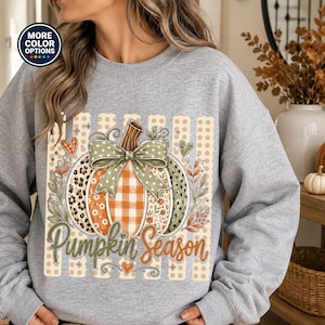 May include: A light grey crewneck sweatshirt featuring a central graphic of a pumpkin with various patterns including leopard print, orange gingham, and floral designs. The pumpkin is topped with a green polka dot bow and the text Pumpkin Season.