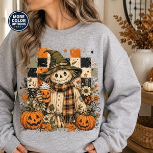 May include: A heather gray crewneck sweatshirt featuring a graphic of a scarecrow wearing a hat and plaid scarf, holding a pumpkin-themed cup. The design includes orange and black checkered patterns, pumpkins, and autumn leaves.