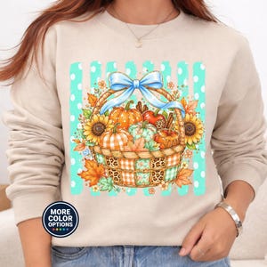 Peut inclure: Un sweat-shirt beige à col rond avec une impression graphique représentant un panier tressé rempli de citrouilles, de tournesols et de feuilles d'automne. Le panier est orné d'un nœud bleu clair sur un fond de rayures verticales bleu sarcelle à pois blancs.