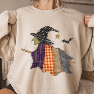 May include: A beige crewneck sweatshirt featuring a central applique of a witch flying on a broomstick. The witch wears a black hat with stars and moons, a green face, and a patchwork cloak made of purple plaid, orange gingham, and gray fabric.